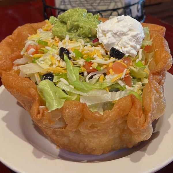 A taco salad in a crispy fried shell, topped with lettuce, cheese, tomatoes, olives, guacamole, and sour cream.