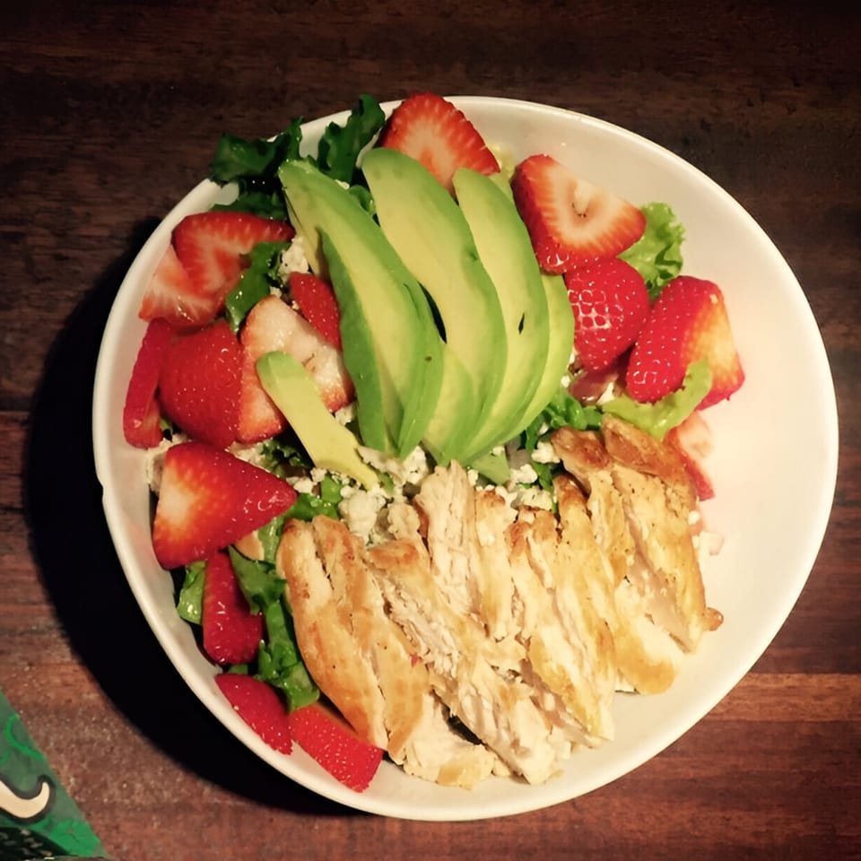 Chicken and strawberry salad with avocado slices and crumbled cheese on a white plate.