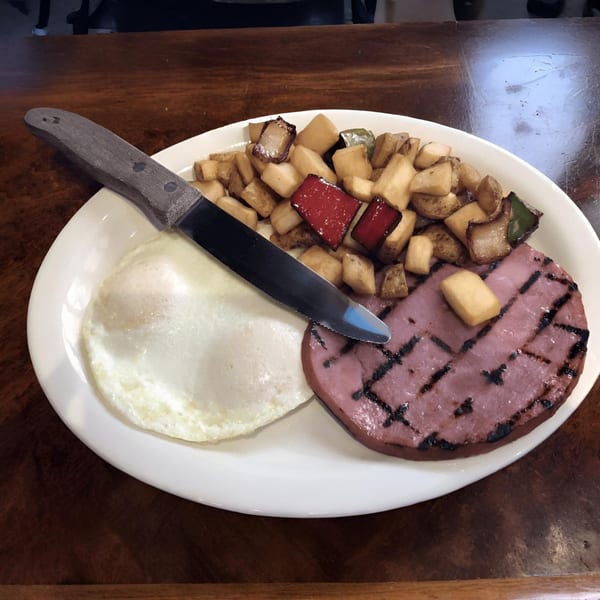 Grilled ham, fried egg, and home fries with peppers on a white plate at Vista Village Pub.