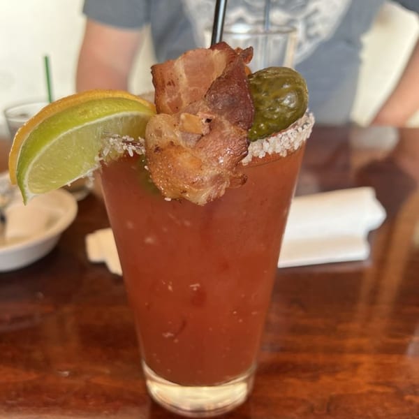 A Bloody Mary cocktail garnished with bacon, a pickle, and a lime wedge, on a wooden table.