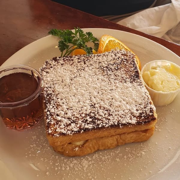 French toast dusted with powdered sugar, served with syrup, butter, orange slices, and parsley.