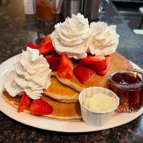 Fluffy pancakes topped with whipped cream, fresh strawberries, and sides of syrup and butter.