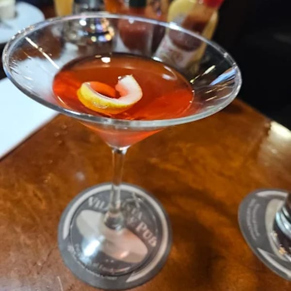 A red cocktail with a lemon twist in a martini glass on a Vista Village Pub coaster.