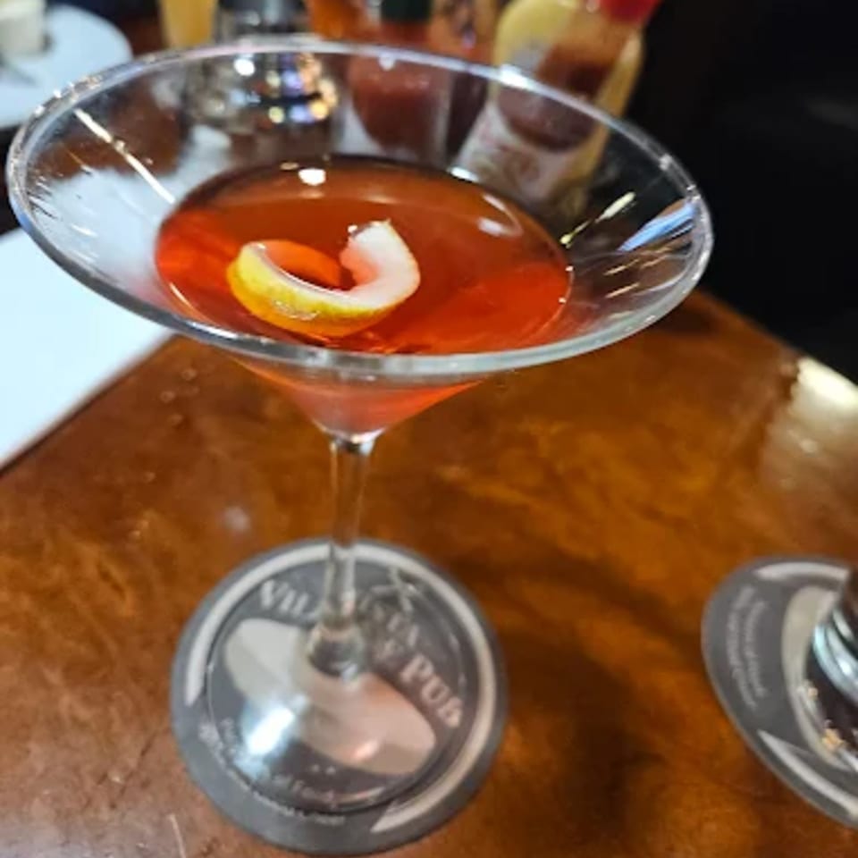 A red cocktail with a lemon twist in a martini glass on a Vista Village Pub coaster.