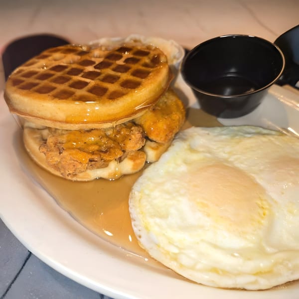 Chicken and waffles with syrup and two fried eggs on a white plate at Vista Village Pub.