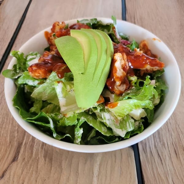 A fresh salad with green lettuce, chicken pieces, avocado slices, and a red sauce in a white bowl.