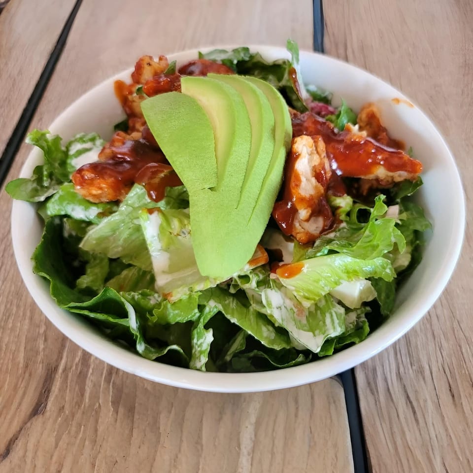 A fresh salad with green lettuce, chicken pieces, avocado slices, and a red sauce in a white bowl.