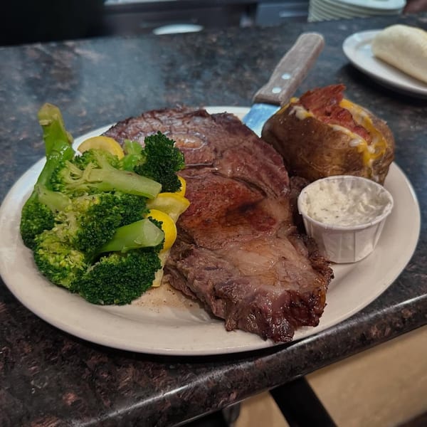 A prime rib dinner with broccoli, yellow squash, a baked potato, and a side of sauce.
