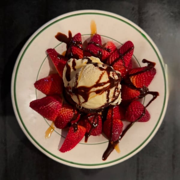 A dessert of vanilla ice cream with chocolate drizzle, surrounded by fresh strawberries on a white plate.